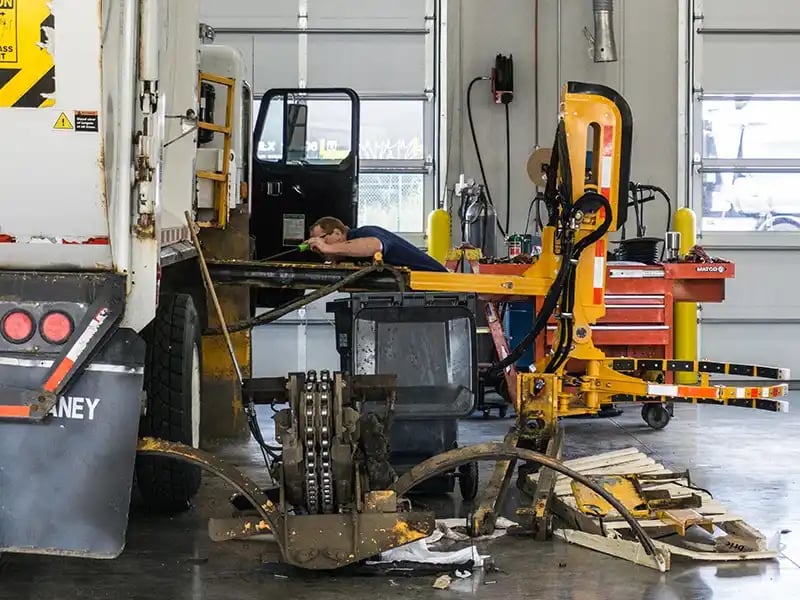 A technician works on the rear section of a refuse truck inside a vehicle maintenance shop with tools.